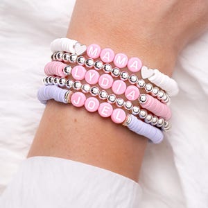 May include: Stack of beaded bracelets in white, pink, and lavender. The bracelets feature silver beads and lettered beads spelling "MAMA", "LYDIA", and "JOEL". Some bracelets have heart-shaped accents. The bracelets are worn on a wrist.
