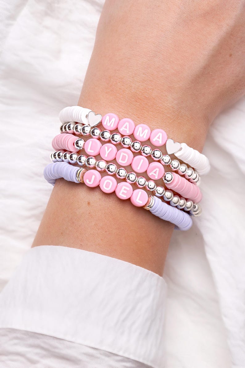 May include: Stack of beaded bracelets in white, pink, and lavender. The bracelets feature silver beads and lettered beads spelling "MAMA", "LYDIA", and "JOEL". Some bracelets have heart-shaped accents. The bracelets are worn on a wrist.
