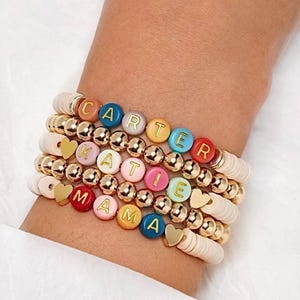 May include: Stacked bracelets with white, gold, and colorful beads. The bracelets spell out "CARTER", "KATIE", and "MAMA" with letter beads. Gold heart charms and gold ball beads add detail to the bracelets.