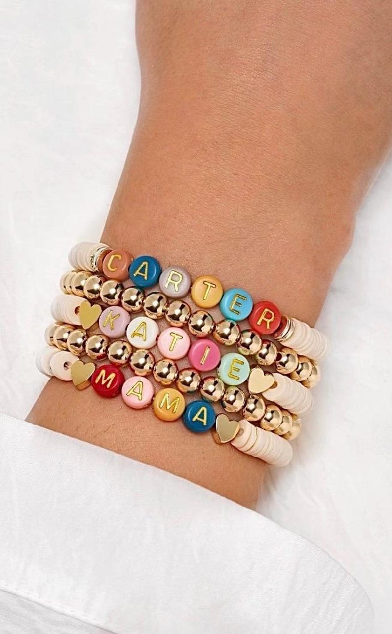 May include: Stacked bracelets with white, gold, and colorful beads. The bracelets spell out "CARTER", "KATIE", and "MAMA" with letter beads. Gold heart charms and gold ball beads add detail to the bracelets.