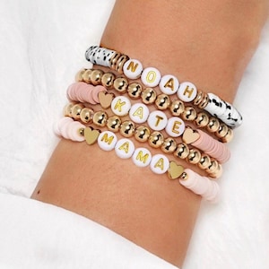 May include: A collection of beaded bracelets. The bracelets feature white letter beads spelling "NOAH", "KATE", and "MAMA", gold beads, pink disc beads, and heart charms. One bracelet has black and white spotted beads.