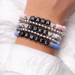 May include: Stack of personalized bracelets with black letter beads spelling "MAMA", "LUKE", and "DANIEL". The bracelets feature silver beads, heart charms, and pastel pink and blue accents. A stylish accessory.