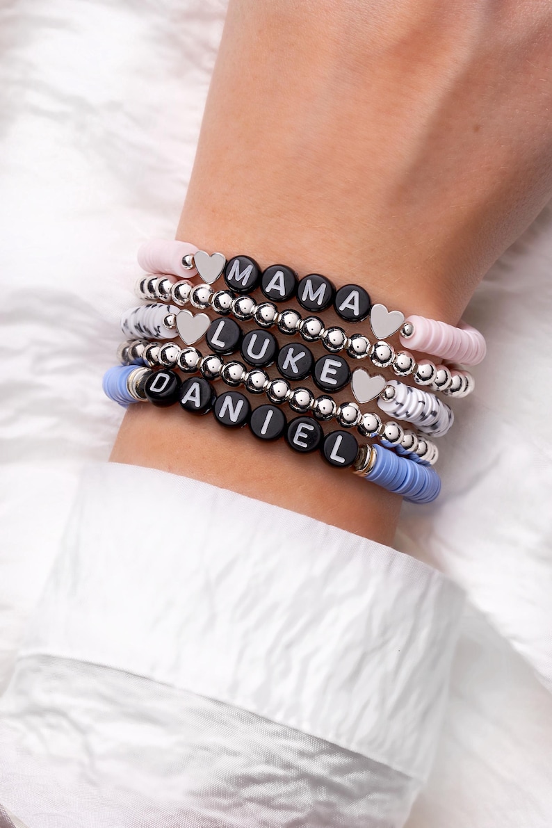 May include: Stack of personalized bracelets with black letter beads spelling "MAMA", "LUKE", and "DANIEL". The bracelets feature silver beads, heart charms, and pastel pink and blue accents. A stylish accessory.