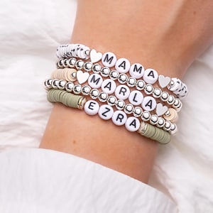 May include: Stack of beaded bracelets with white letter beads spelling "MAMA", "MARLA", and "EZRA". The bracelets include silver beads, heart charms, and a mix of white, beige, and olive green disc beads. The bracelets are stacked on a wrist.
