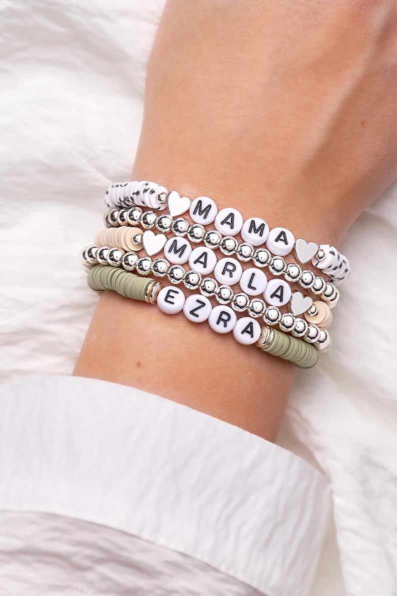 May include: Stack of beaded bracelets with white letter beads spelling "MAMA", "MARLA", and "EZRA". The bracelets include silver beads, heart charms, and a mix of white, beige, and olive green disc beads. The bracelets are stacked on a wrist.