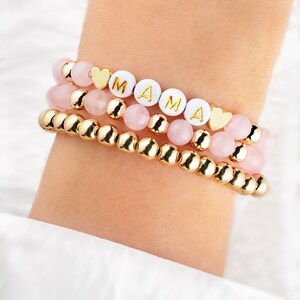 May include: Three beaded bracelets stacked on a wrist. The top bracelet has white beads spelling "MAMA" with gold heart accents. The middle bracelet has pink beads, and the bottom bracelet has gold beads. The bracelets are a mix of gold and pink.