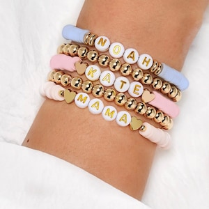 May include: A selection of beaded bracelets in pastel shades of blue, pink, and white. Each bracelet features gold-coloured beads and charms, with lettered beads spelling out names. The bracelets are worn on a wrist.