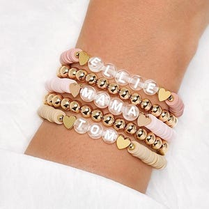 May include: A collection of personalised bracelets. The bracelets feature white letter beads spelling "ELLIE", "MAMA", and "TOM". They also include gold beads, heart charms, and pastel-coloured disc beads. The bracelets are stacked on a wrist.