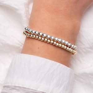 May include: Two beaded bracelets, one silver and one gold, are stacked on a wrist. The bracelets are made of small, round beads. The wrist is resting on a white shirt.