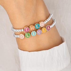 May include: Two stretch bracelets made of small silver-toned metallic beads. Each bracelet features round, gold-rimmed enamel charms in various pastel colours. The top bracelet spells THEO and the bottom bracelet spells OLIVIA in colourful letters.