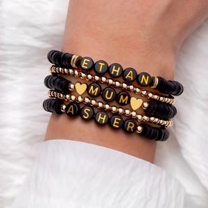 May include: Stack of black beaded bracelets with gold accents and letter beads spelling out names: ETHAN, MUM, and ASHER. Each bracelet has a different design, with some featuring gold heart charms.