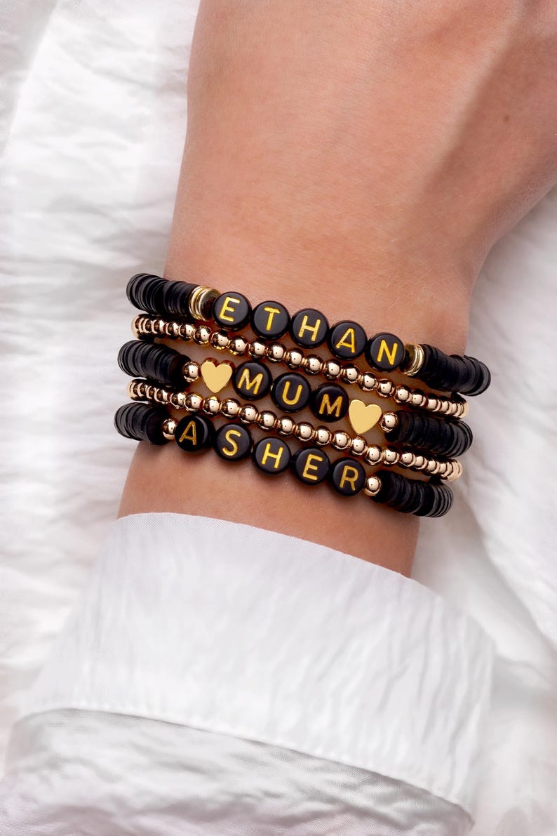 May include: Stack of black beaded bracelets with gold accents and letter beads spelling out names: ETHAN, MUM, and ASHER. Each bracelet has a different design, with some featuring gold heart charms.