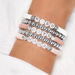 May include: A collection of personalised bracelets spelling "NOAH", "KATE", and "MAMA". The bracelets feature silver beads, pink disc beads, and heart charms. The bracelets are worn on a wrist.