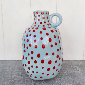 May include: A light blue ceramic vase with a red polka dot pattern and a handle. The vase is about 10 inches tall.