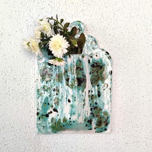 May include: A ceramic wall vase with a rectangular shape and a handle at the top. The vase is glazed in shades of turquoise, white, and green, with a speckled pattern. White flowers and green foliage are arranged in the vase.