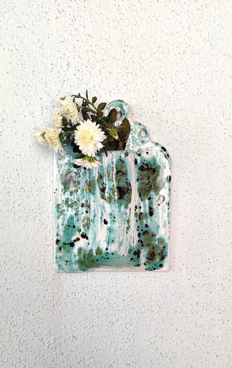 May include: A ceramic wall vase with a rectangular shape and a handle at the top. The vase is glazed in shades of turquoise, white, and green, with a speckled pattern. White flowers and green foliage are arranged in the vase.