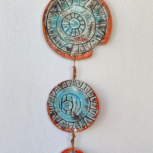 Op de afbeelding: Handgemaakte keramische wandhanger met drie cirkelvormige, getextureerde stukken. Elk stuk heeft een blauw midden met een geometrisch ontwerp, omgeven door een rood-oranje rand. De stukken zijn verbonden door een natuurlijke vezelkoord.