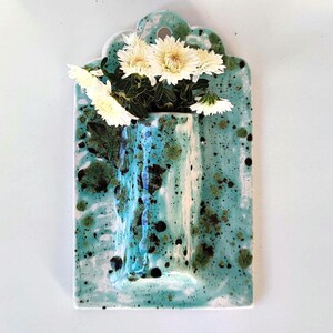 May include: A rectangular ceramic wall vase with a turquoise glaze and dark green and black speckles. The vase holds white chrysanthemums. The top of the vase has a scalloped edge.