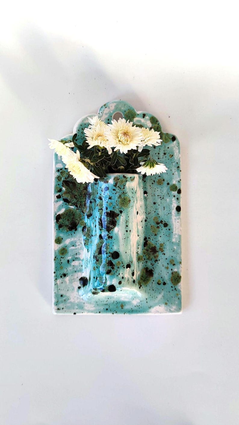 May include: A rectangular ceramic wall vase with a turquoise glaze and dark green and black speckles. The vase holds white chrysanthemums. The top of the vase has a scalloped edge.
