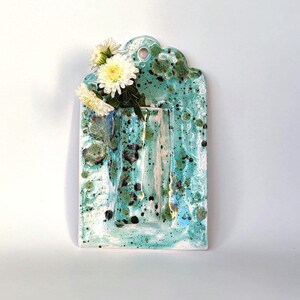 May include: A ceramic wall vase with a turquoise and white speckled glaze. The vase has a rectangular shape with a scalloped top and a hole for hanging. White and yellow flowers are arranged in the vase.