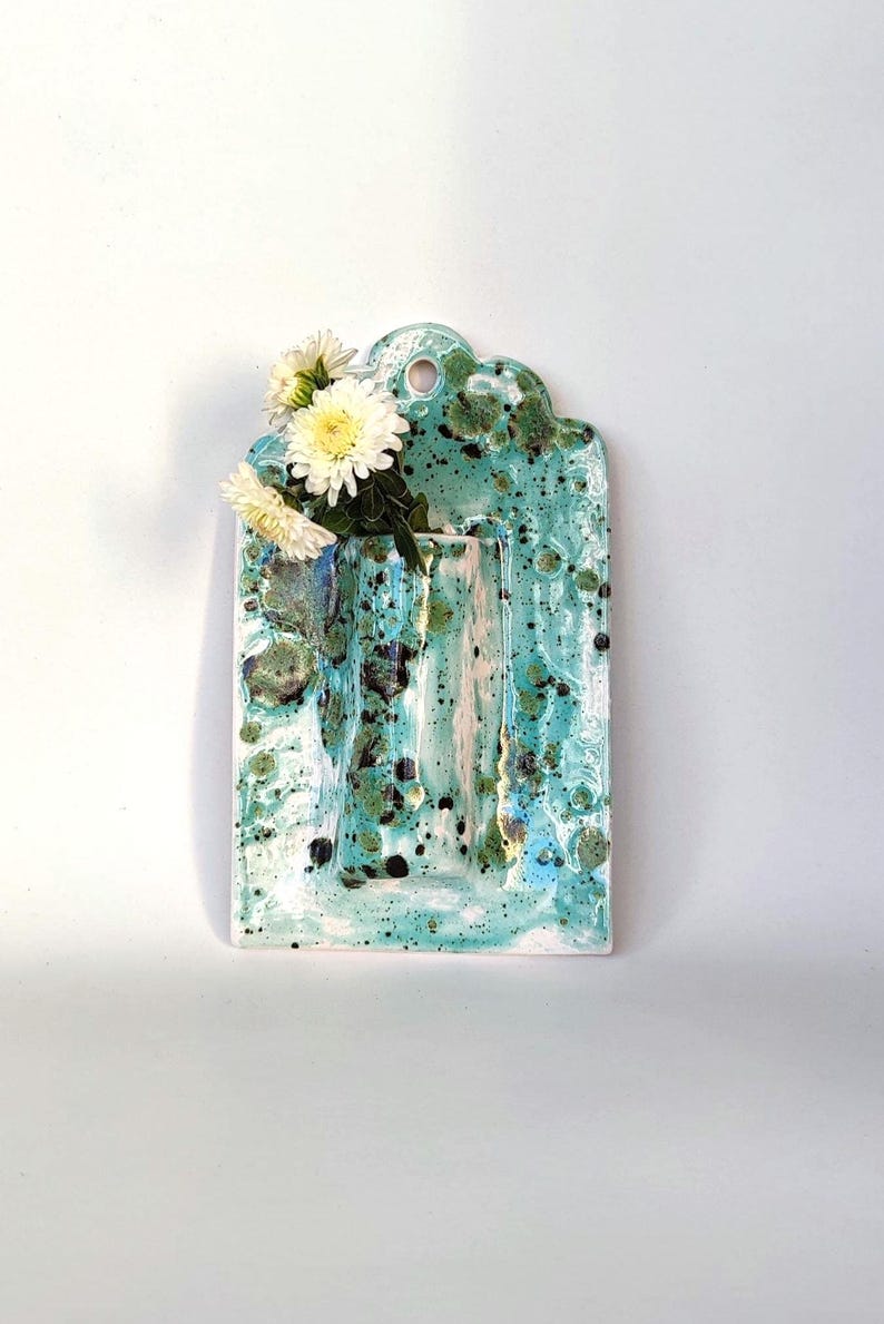May include: A ceramic wall vase with a turquoise and white speckled glaze. The vase has a rectangular shape with a scalloped top and a hole for hanging. White and yellow flowers are arranged in the vase.