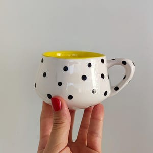 Può includere: Una tazza in ceramica bianca con pois neri e interno giallo. La tazza ha una forma arrotondata e un manico. La tazza è tenuta in mano. È un articolo fatto a mano.