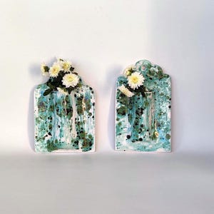May include: Two ceramic wall vases with a turquoise and white speckled glaze. Each vase has a rectangular shape with a scalloped top and is filled with white flowers. The vases are displayed against a white background.