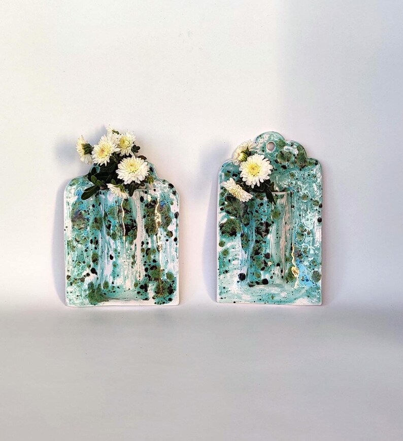 May include: Two ceramic wall vases with a turquoise and white speckled glaze. Each vase has a rectangular shape with a scalloped top and is filled with white flowers. The vases are displayed against a white background.