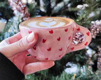 May include: A pink ceramic coffee cup adorned with red heart shapes, filled with a latte featuring heart-shaped foam art. The cup is held in a hand, with a blurred background of a Christmas tree.