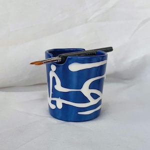 Puede incluir: Un portacepillos de cerámica azul con diseños abstractos blancos. Dos pinceles están en el portacepillos.