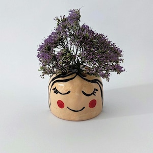 May include: A small, round, beige ceramic planter shaped like a face with closed eyes, long black eyelashes, a simple curved mouth, and two red circular cheeks. The planter is filled with a cluster of small, vibrant purple flowers.