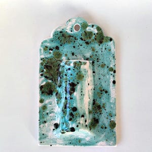 May include: A ceramic wall pocket in a rectangular shape with a scalloped top edge. The piece is glazed in a turquoise color with dark green and black speckles. It has a built-in vase for displaying flowers.