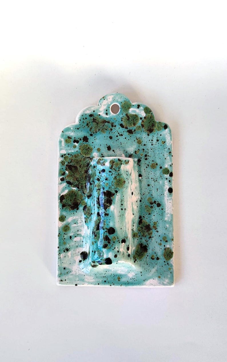May include: A ceramic wall pocket in a rectangular shape with a scalloped top edge. The piece is glazed in a turquoise color with dark green and black speckles. It has a built-in vase for displaying flowers.