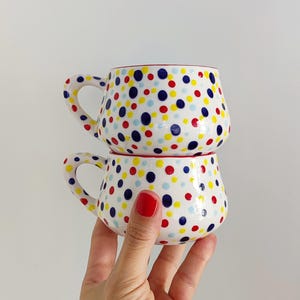 Puede incluir: Dos tazas de cerámica blanca apiladas, cada una adornada con un vibrante patrón de lunares rojos, amarillos y azules. Las tazas tienen cuerpos redondeados, bordes rojos y asas curvas. Las tazas son sostenidas por una mano.
