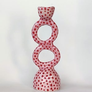 Puede incluir: Un portavelas de cerámica rosa con un diseño único. El portavelas presenta una serie de formas circulares conectadas, cada una adornada con lunares rojos. La base y la parte superior también tienen el mismo patrón de lunares.