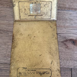 Könnte beinhalten: Ein goldfarbener, alter ewiger Kalender aus Metall mit einem rechteckigen oberen Teil, der den Text H. Ells Oil Co. Kaiser-Frazer Agency Hebron, Nebraska und einen Kalender enthält. Der untere Teil zeigt Anweisungen zum Einstellen des Kalenders.