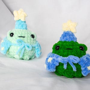 May include: Two crocheted frog plushies wearing blue and yellow party hats and scarves. The frogs are green and light green.