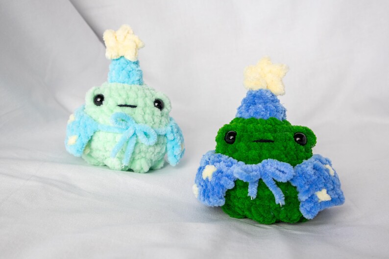 May include: Two crocheted frog plushies wearing blue and yellow party hats and scarves. The frogs are green and light green.