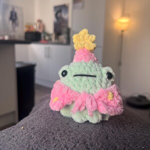 May include: A green crocheted frog wearing a pink and yellow party hat with a yellow star on top. The frog is sitting on a gray fuzzy surface.