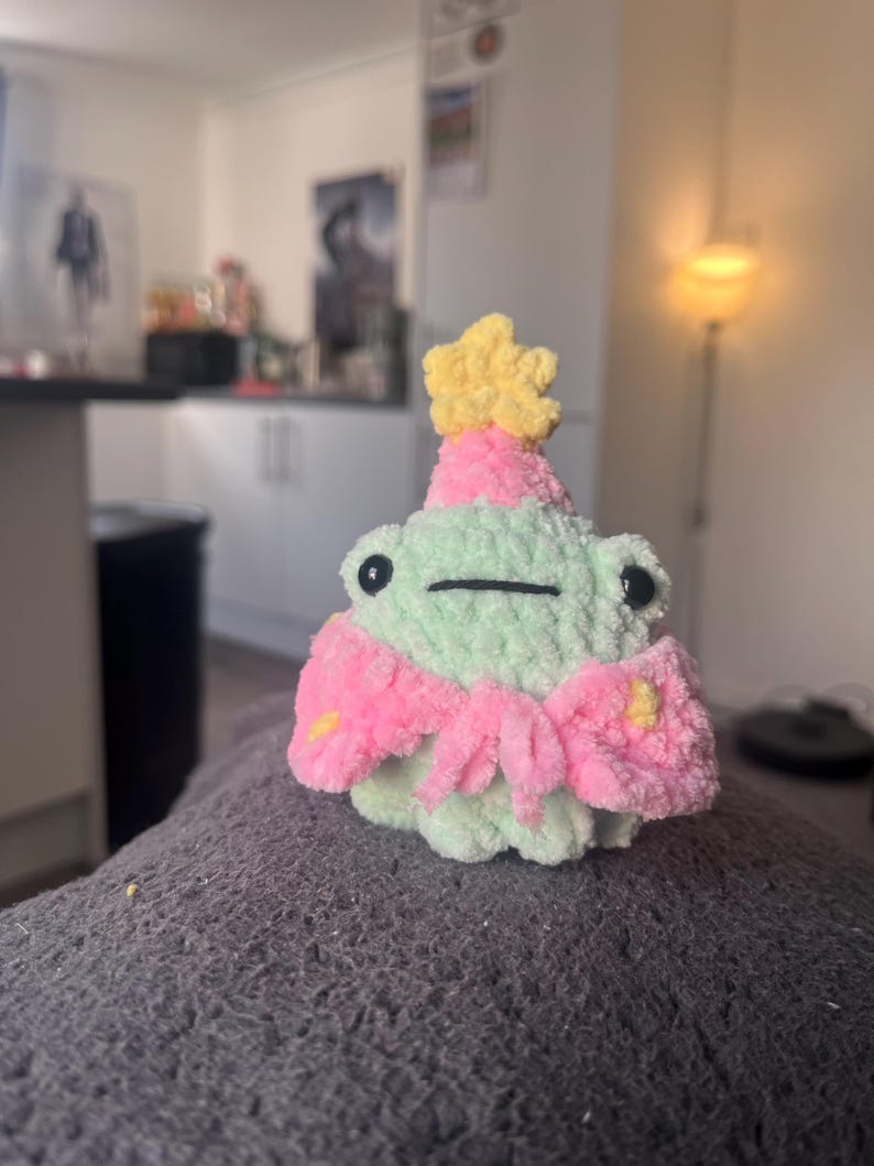 May include: A green crocheted frog wearing a pink and yellow party hat with a yellow star on top. The frog is sitting on a gray fuzzy surface.