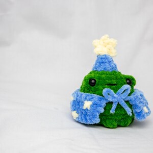 May include: A green crocheted frog wearing a blue and white crocheted hat and a blue crocheted scarf with a white star on it.