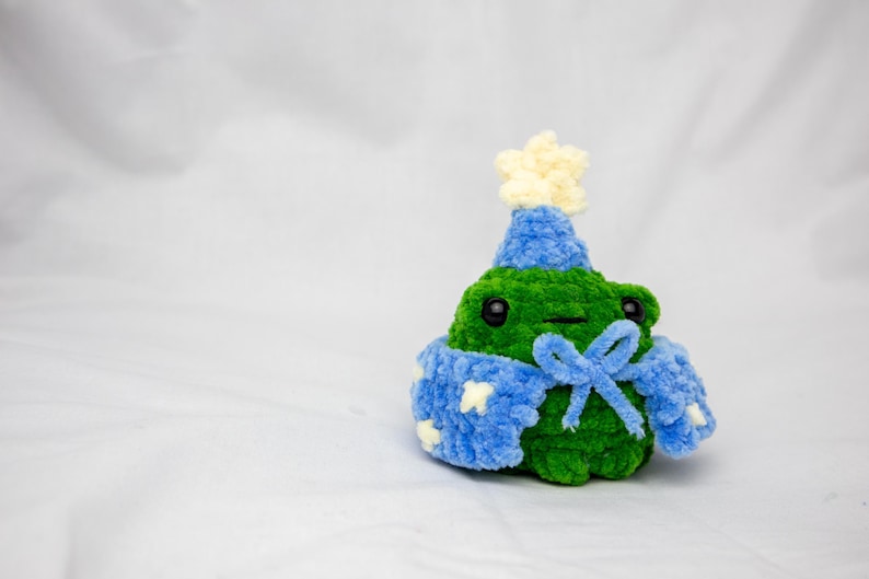 May include: A green crocheted frog wearing a blue and white crocheted hat and a blue crocheted scarf with a white star on it.