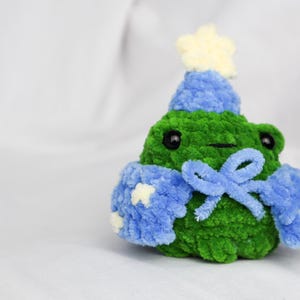 May include: A green crocheted frog wearing a blue crocheted hat with a white star on top and a blue crocheted scarf with a bow.