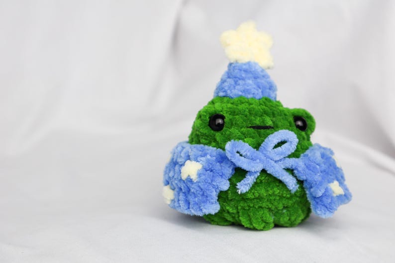 May include: A green crocheted frog wearing a blue crocheted hat with a white star on top and a blue crocheted scarf with a bow.