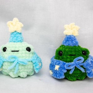 May include: Two crocheted frog plushies wearing blue and white hats and scarves. The frog on the left is light green and the frog on the right is green.