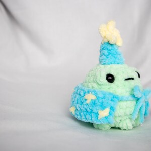 May include: A green crocheted plush toy wearing a blue and yellow crocheted hat and a blue crocheted scarf with yellow stars.