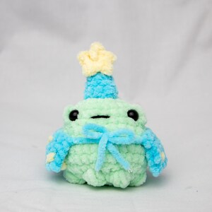 May include: A green crocheted frog wearing a blue and yellow crocheted hat and a blue crocheted scarf with a yellow star on top.