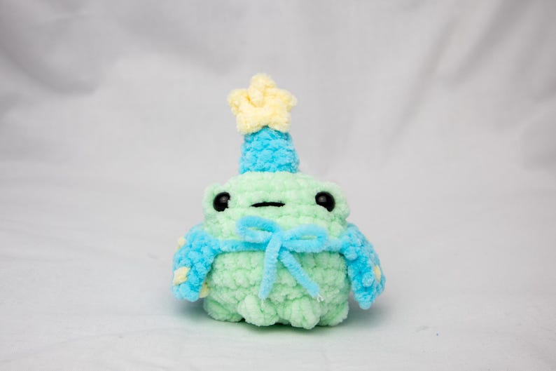 May include: A green crocheted frog wearing a blue and yellow crocheted hat and a blue crocheted scarf with a yellow star on top.