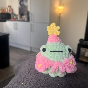 May include: A green crocheted frog wearing a pink and yellow crocheted hat and a pink crocheted scarf with yellow stars.
