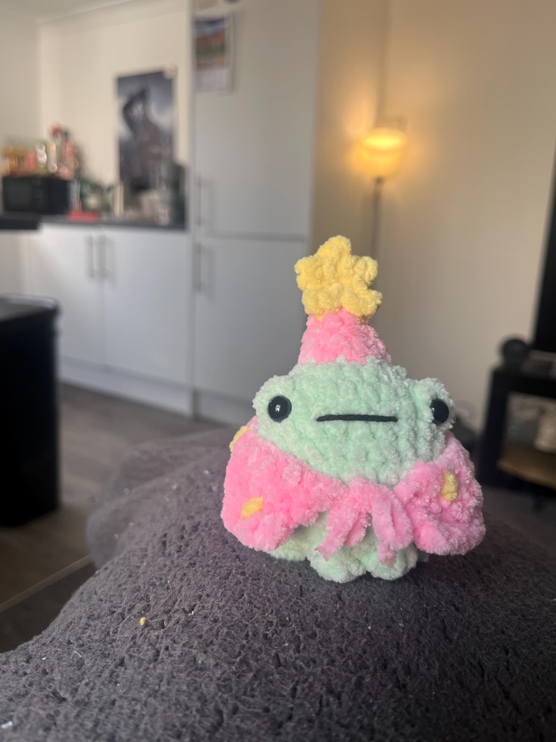 May include: A green crocheted frog wearing a pink and yellow crocheted hat and a pink crocheted scarf with yellow stars.